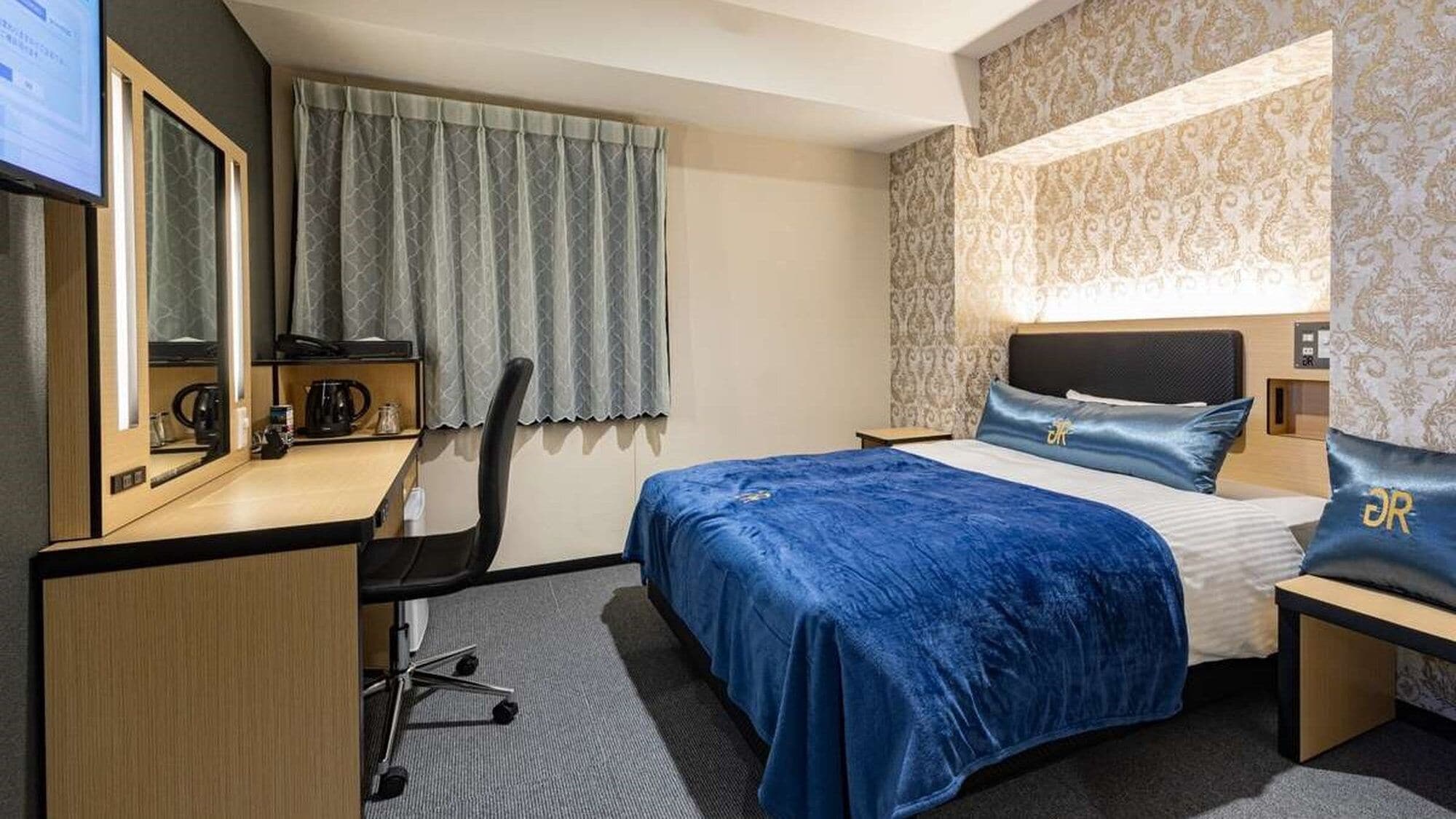 Une chambre d’hôtel comprenant un lit, un bureau, une chaise, une fenêtre avec des rideaux et un mur orné d’un motif décoratif.