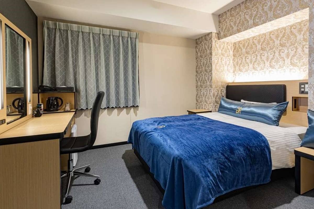 Ein Hotelzimmer mit Bett, Schreibtisch, Stuhl, einem Fenster mit Vorhängen und einer Wand mit einem gemusterten Design.