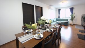 A dining area with a wooden table set for four, surrounded by chairs. There are vases with flowers on the table, and a sofa is visible in the background. - Sakura34 (Sapporo)