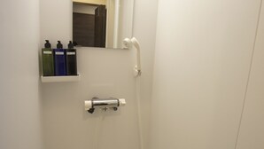 Une petite salle de bain blanche, équipée d’une douche, d’un miroir et d’une étagère où sont rangés les articles de toilette.