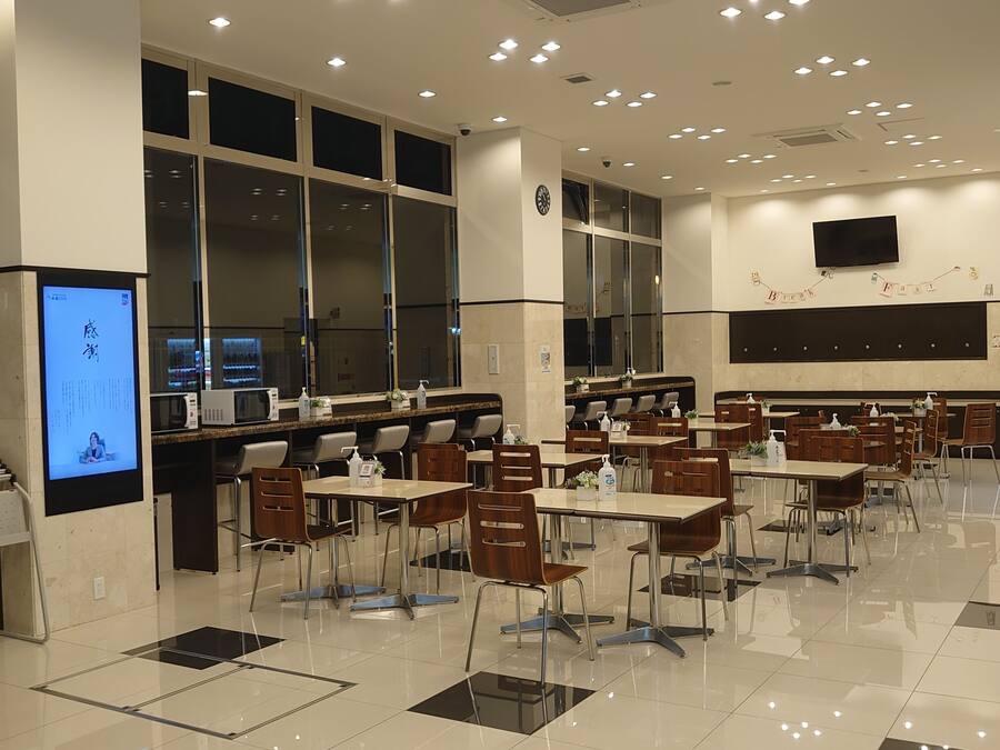 A modern cafeteria with multiple tables and chairs, a large clock, and a television mounted on the wall.