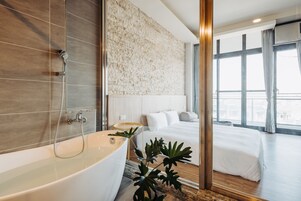 Habitación de hotel moderna con una bañera grande, una cama con ropa blanca, un ventanal con cortinas y una pared con textura.