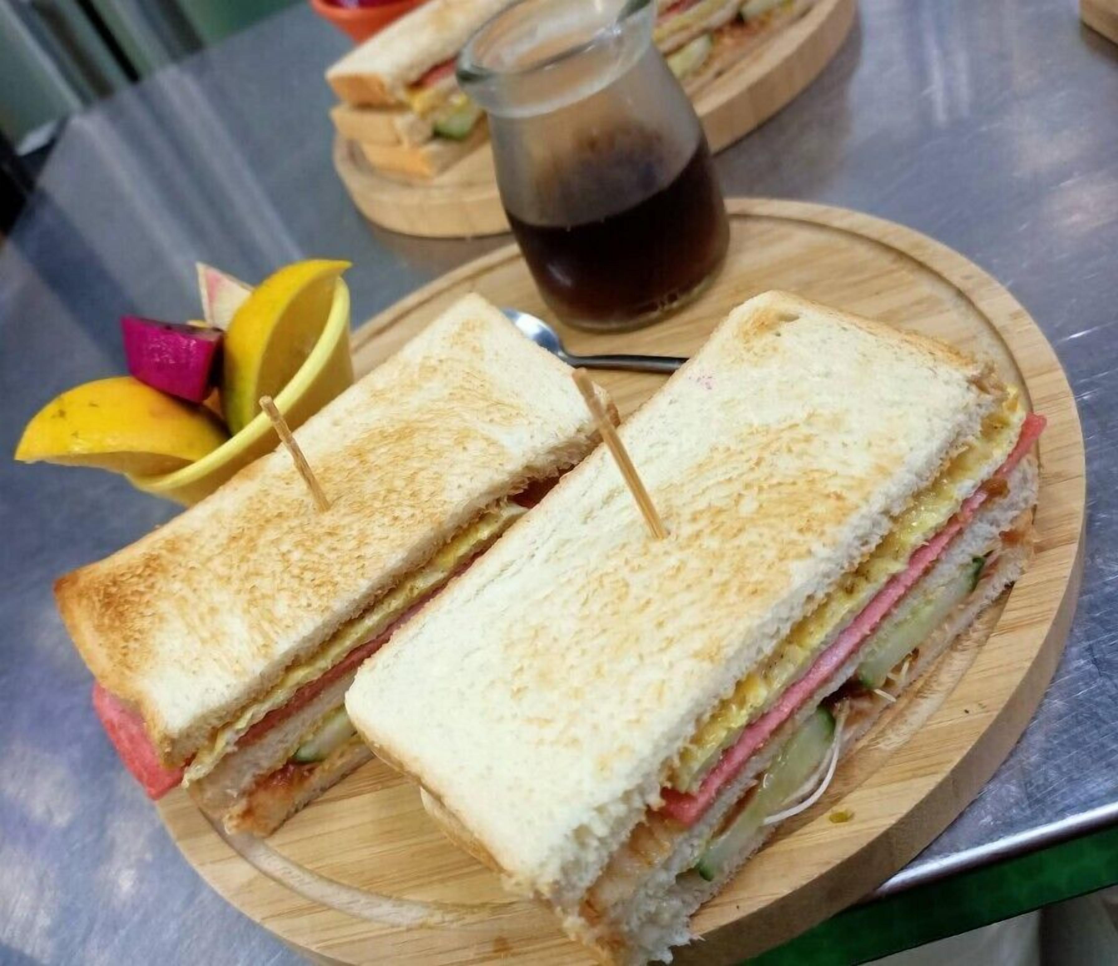 A sandwich cut in half on a wooden board, with a bottle of soda and fruit slices in the background.