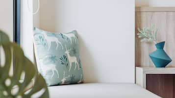 A neatly arranged room with a teal pillow featuring a deer pattern, a wooden side table with a blue vase, and a large green plant in the foreground.