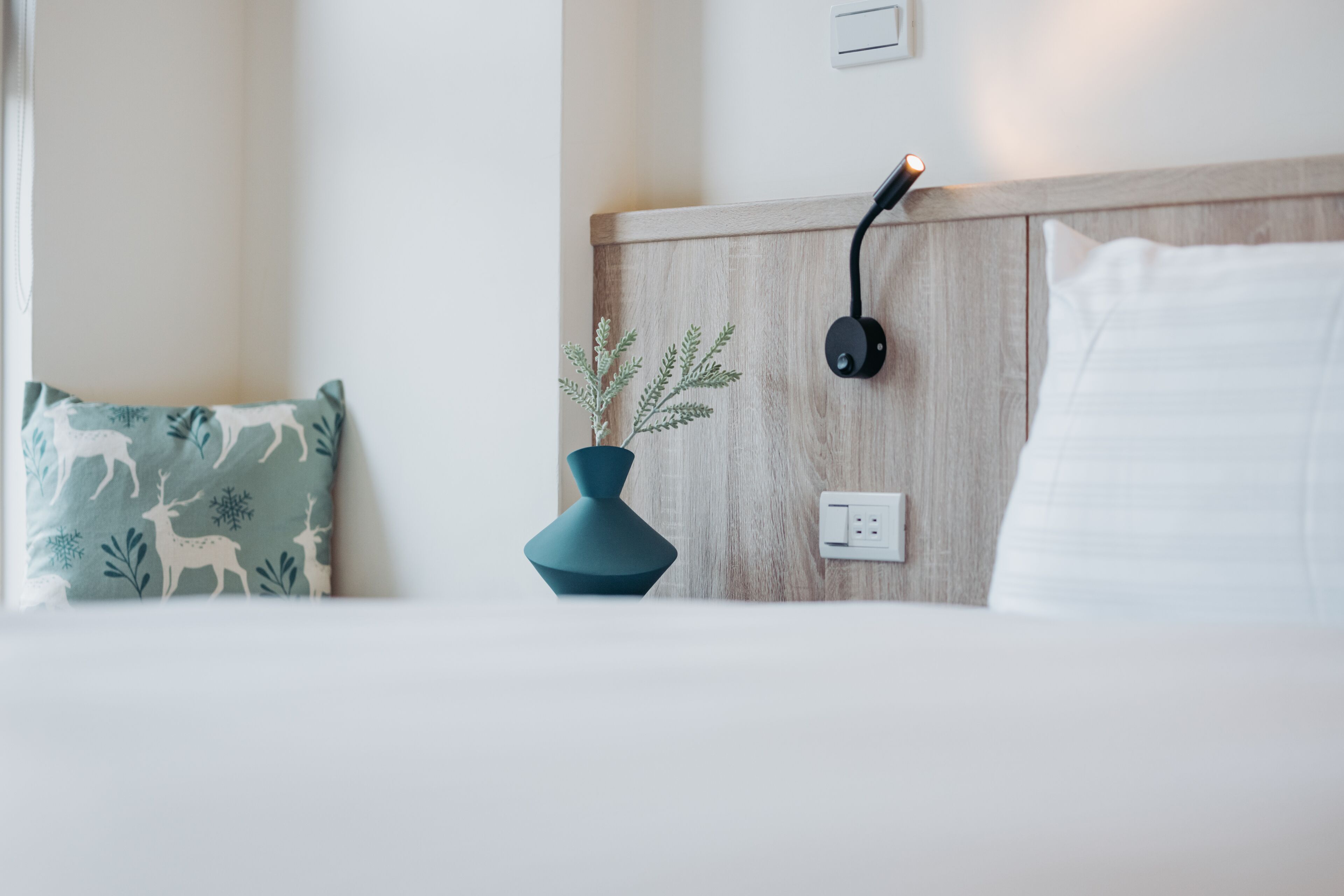 A neatly made bed with white linens, a teal pillow with a deer pattern, a blue vase with a plant, and a wall-mounted charger.