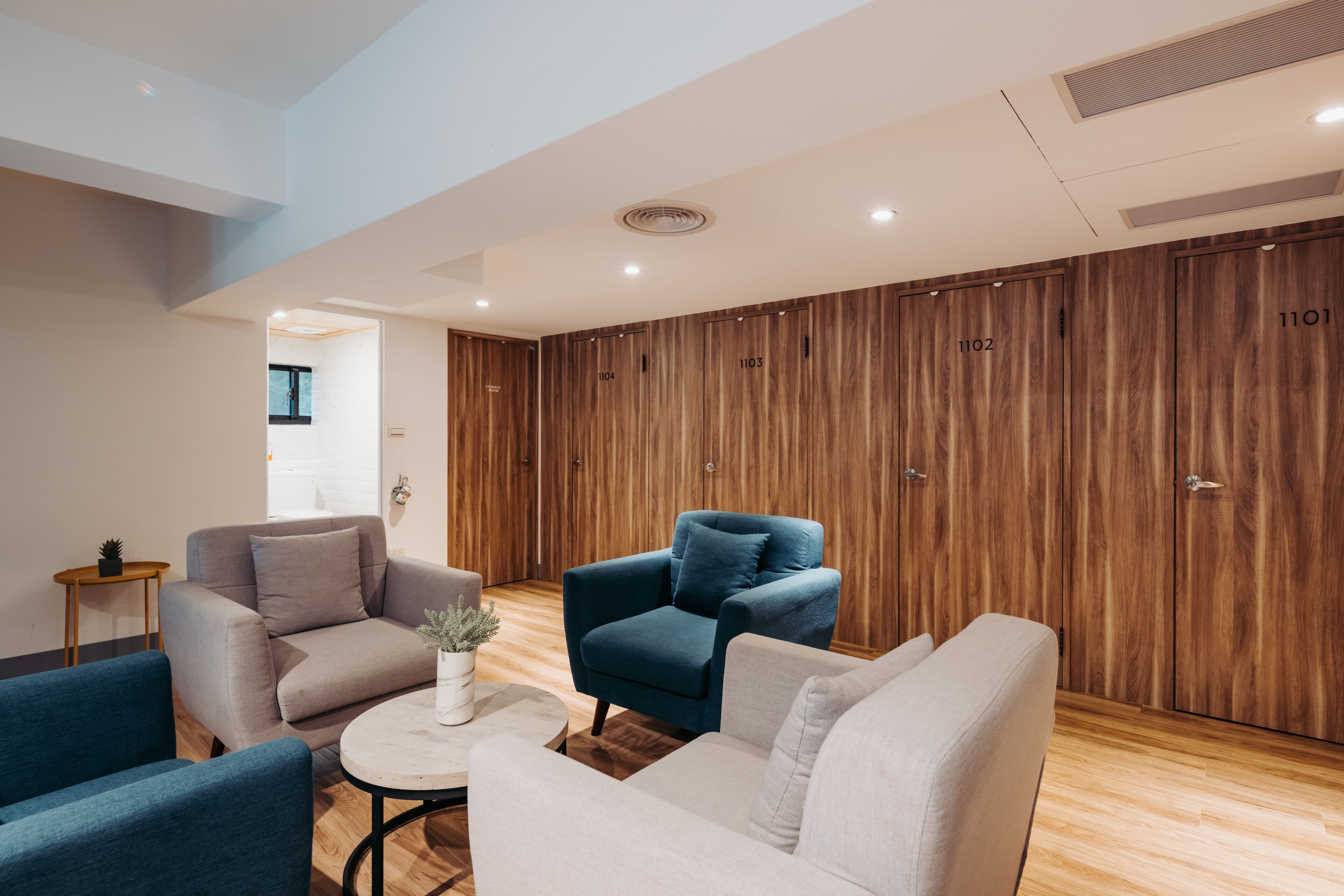 Un moderno vestíbulo de hotel con taquillas de madera, un sofá gris, un sillón azul y una mesa de centro redonda.