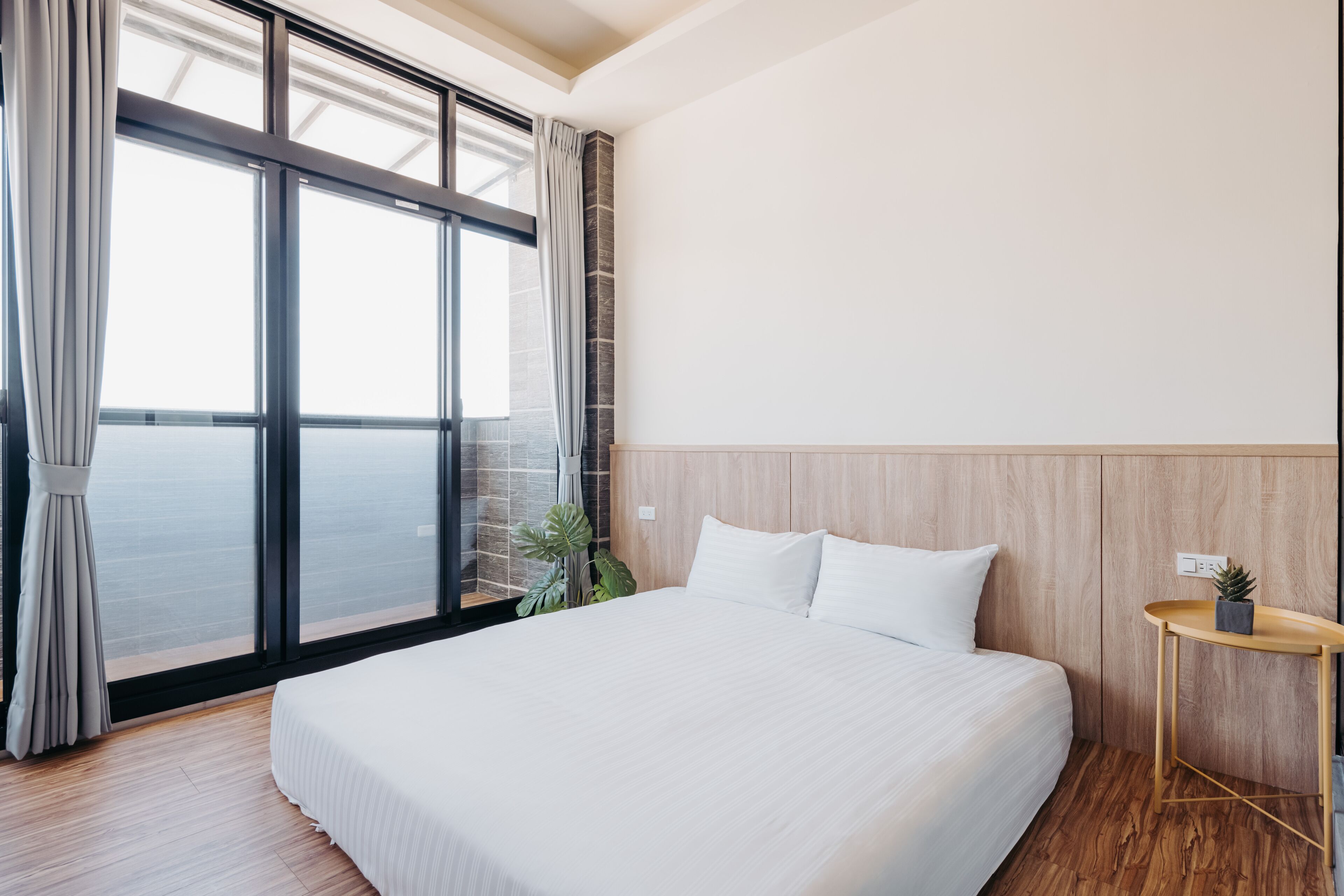 A modern hotel room with a large bed, a wooden headboard, a small table with a plant, and a view of the sea through floor-to-ceiling windows.