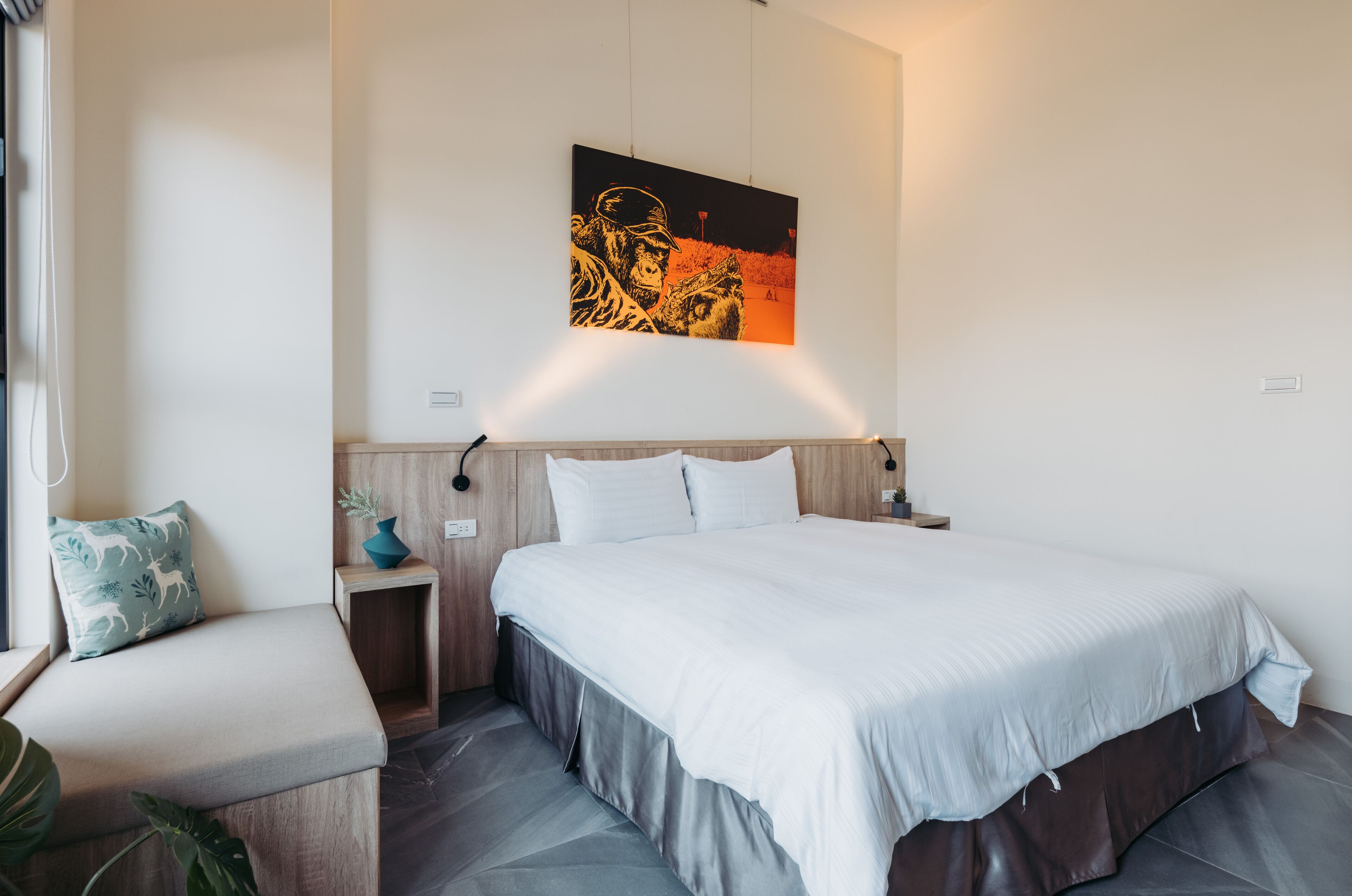 A modern hotel room with a large bed, a bedside table, a small seating area, and a wall-mounted artwork.