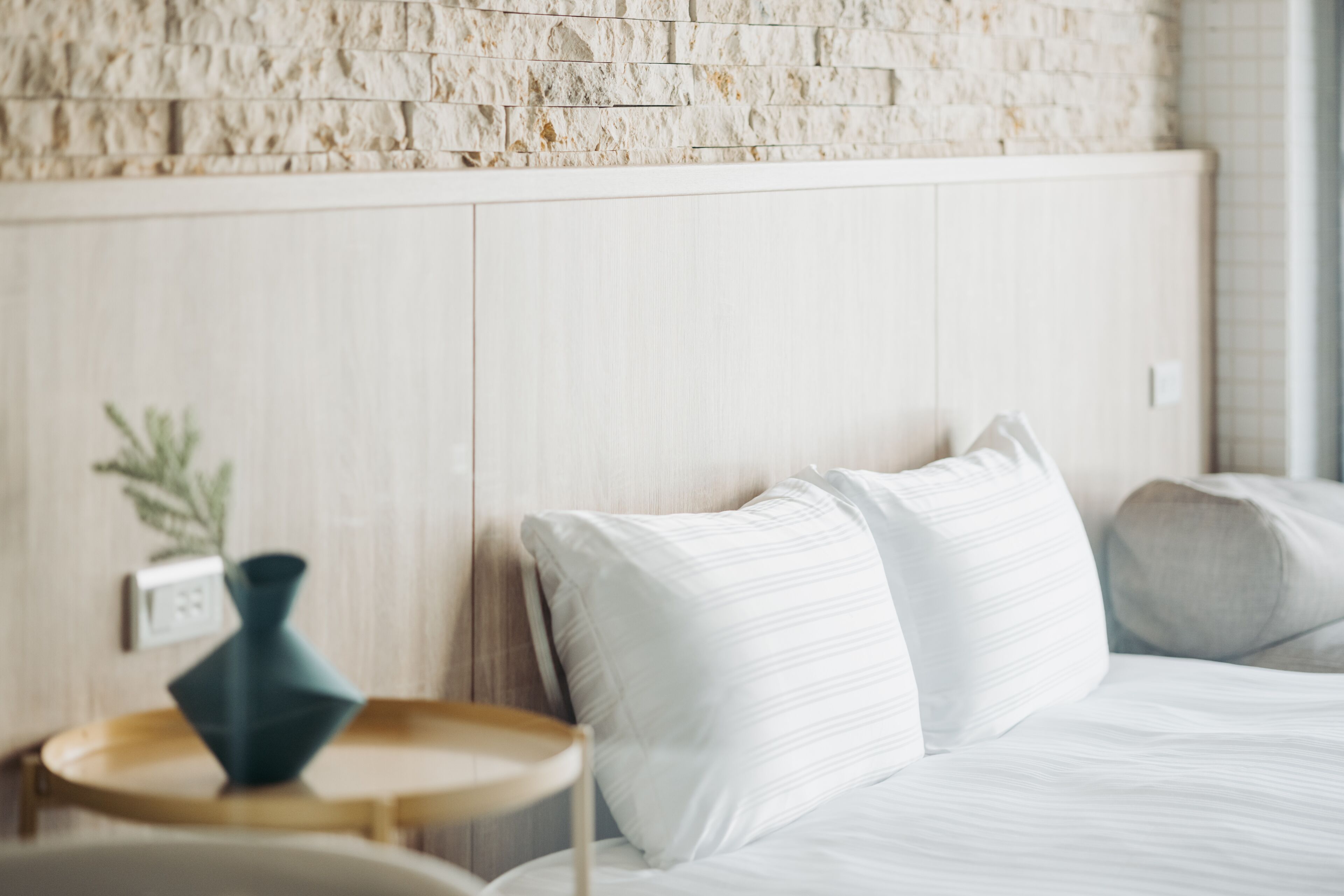 Um quarto com parede de pedra, cama com roupa de cama branca e uma pequena mesa redonda com um vaso.