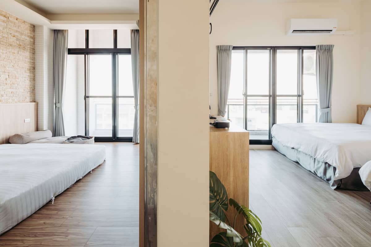 Ein modernes Hotelzimmer mit zwei Betten, großen Fenstern mit Vorhängen, Holzboden und einer Klimaanlage.