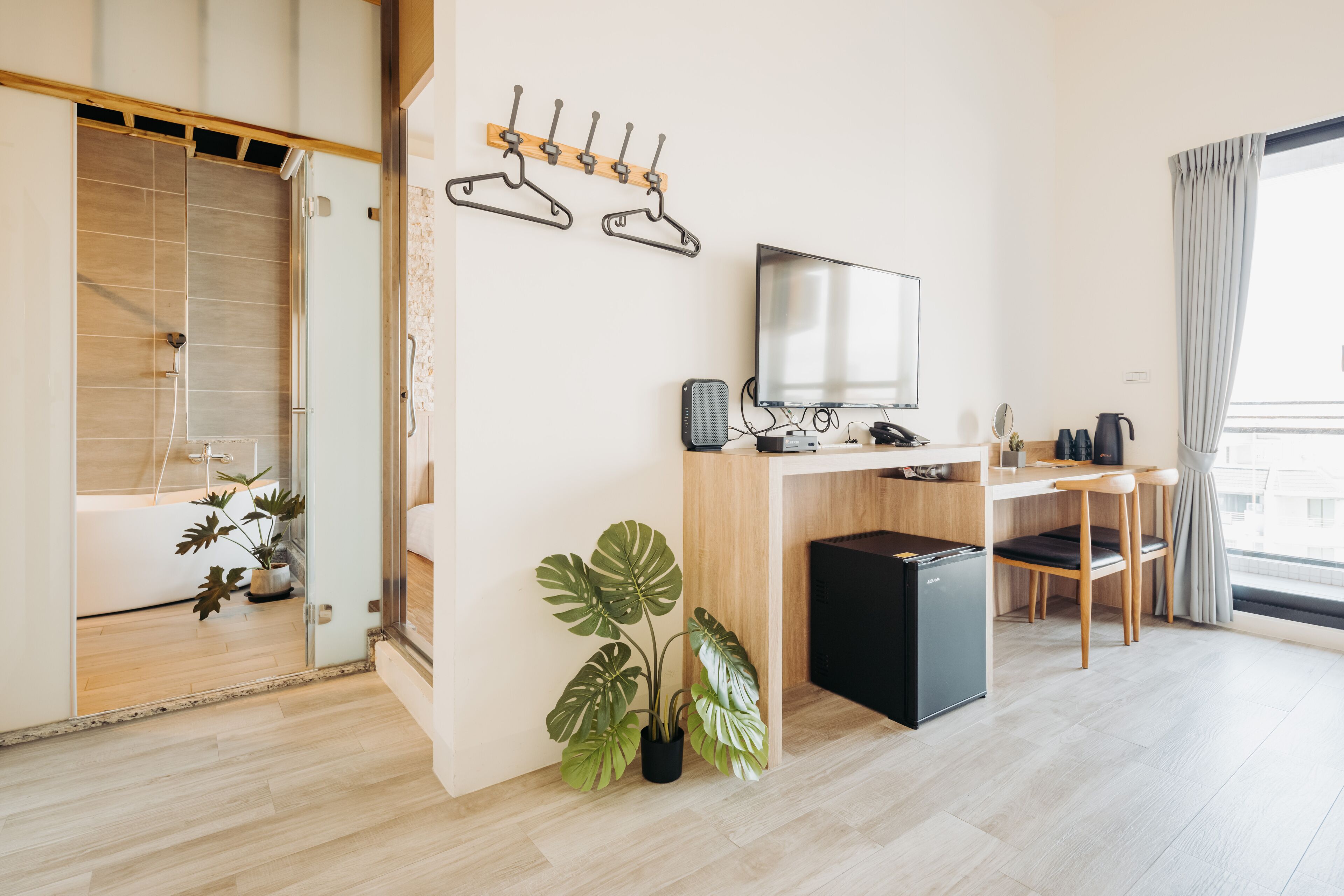 Una habitación de hotel moderna con un escritorio de madera, un televisor de pantalla plana, un refrigerador pequeño, una planta en maceta y un área de ducha.