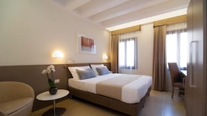A hotel room with a bed, a small round table with a flower, a chair, and a desk with a chair. - Leone (Venice)