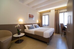 A hotel room with a bed, a small round table with a flower, a chair, and a desk with a chair. - Leone (Venice)