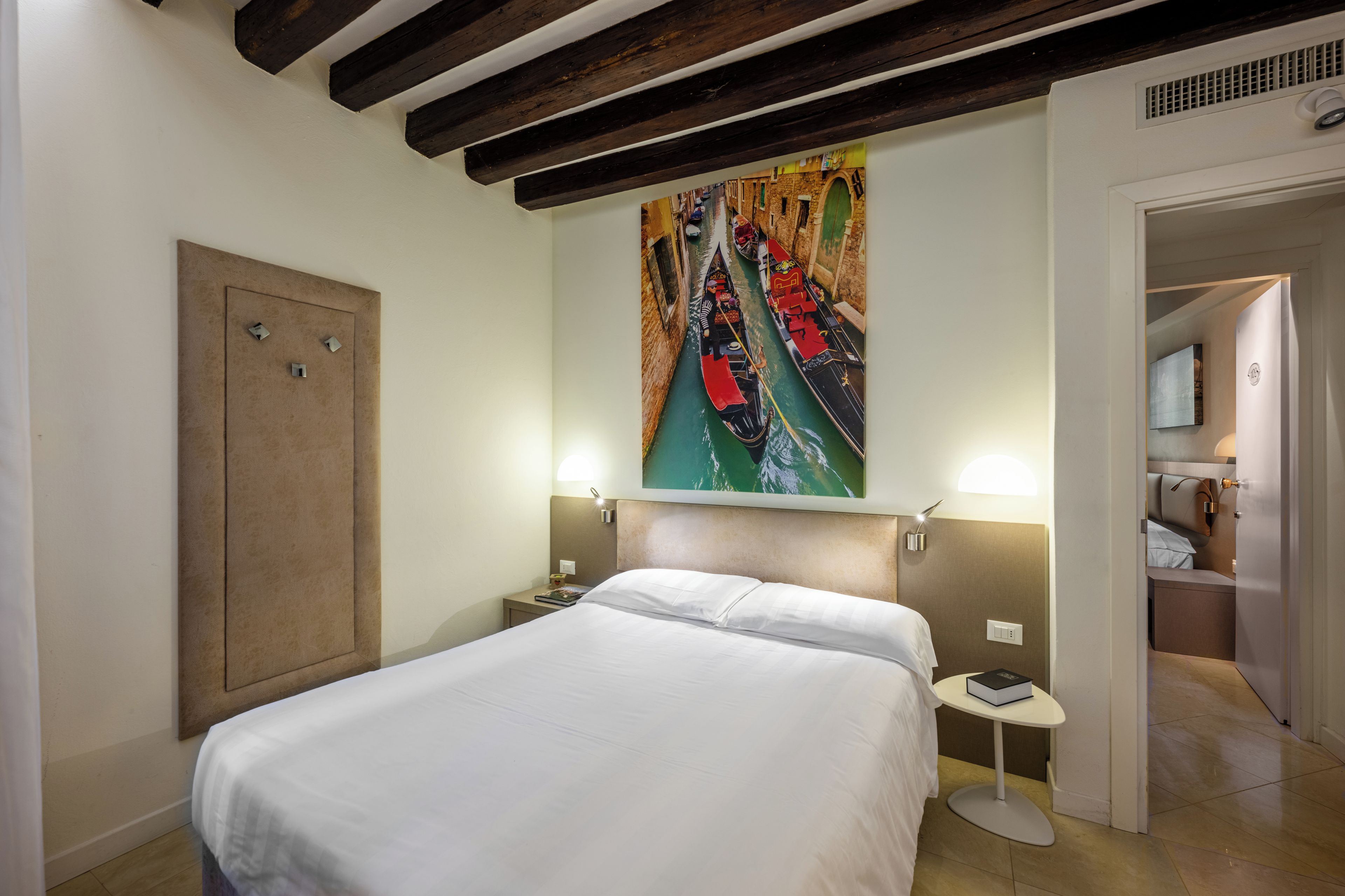 A hotel room with a bed, a painting of a canal, a nightstand, a bedside table, and a door leading to a bathroom.