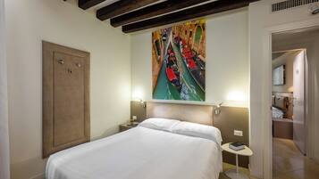 A hotel room with a bed, a painting of a canal, a nightstand, a bedside table, and a door leading to a bathroom.