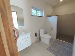 Une salle de bain avec un lavabo, des toilettes et une douche, le tout de couleur blanche. Il y a une fenêtre au-dessus des toilettes et un miroir au-dessus du lavabo.