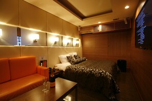 Habitación de hotel con cama, sofá, una mesa con una botella y vasos, y luces fijadas a la pared.