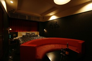 A modern hotel room with a curved red sofa, a small table with a bottle and glasses, and a bed with patterned bedding. - Hotel Pasion - Adults Only		 (Chiba)