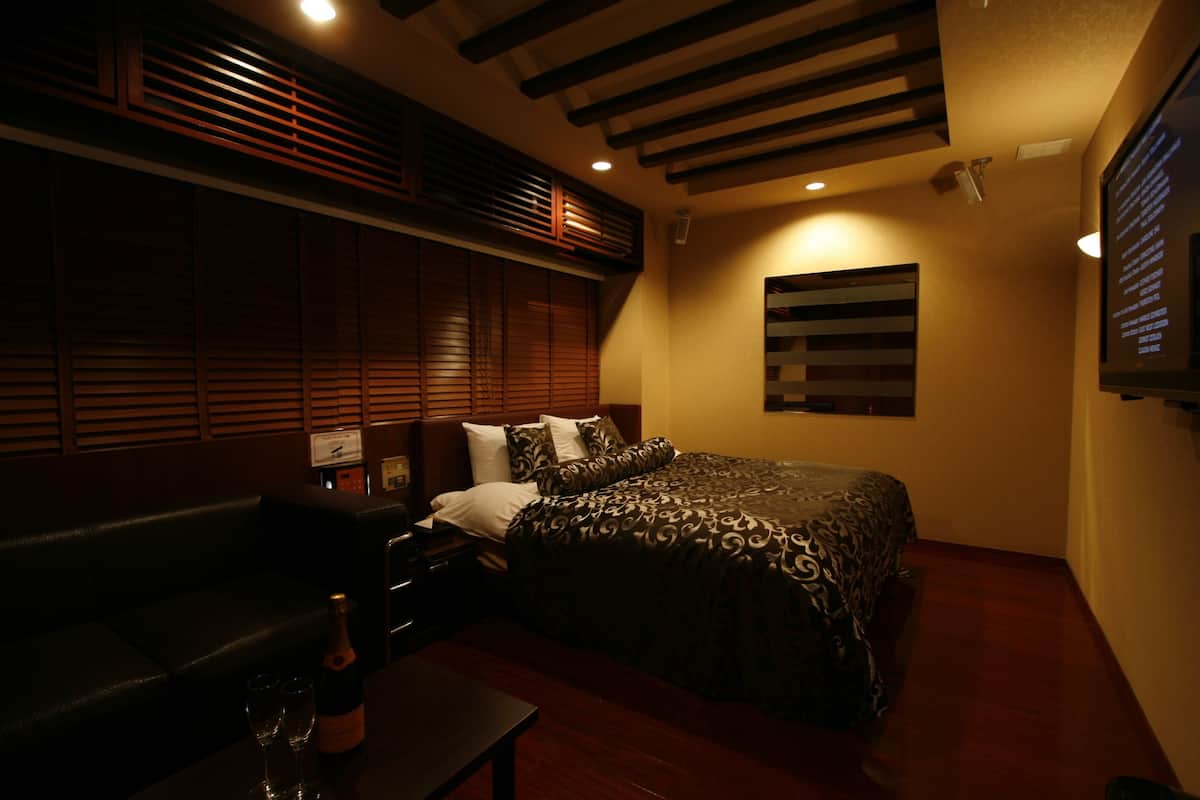 A dimly lit room with a bed, a small table with a bottle and glasses, and a dark leather couch.