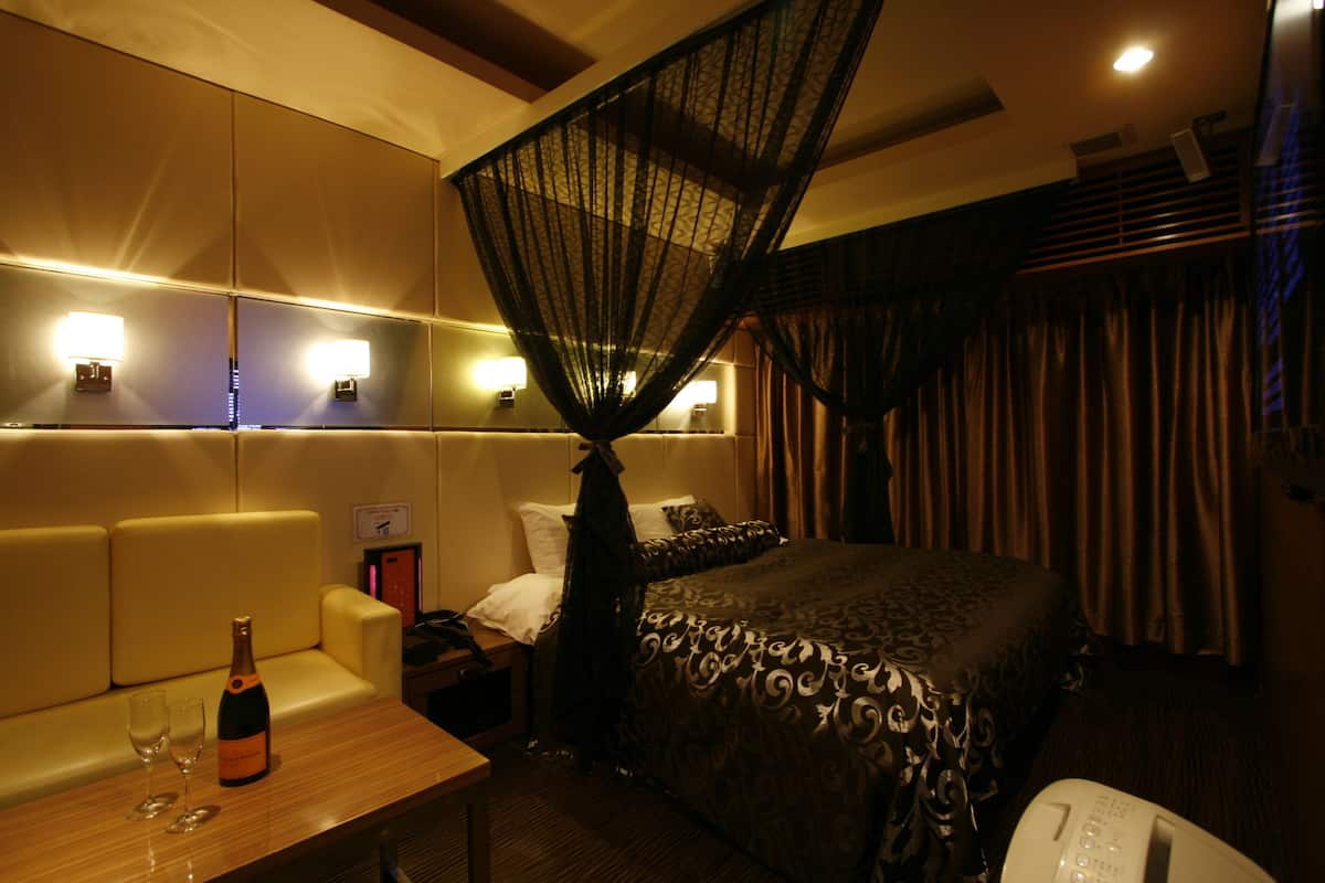 A hotel room with a bed, a bedside table with a bottle and glasses, a nightstand, a canopy bed, and a wall-mounted lamp.