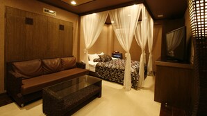 Quarto de hotel com sofá de couro marrom, mesa de centro de vidro, cama com dossel e televisão sobre um móvel de madeira.