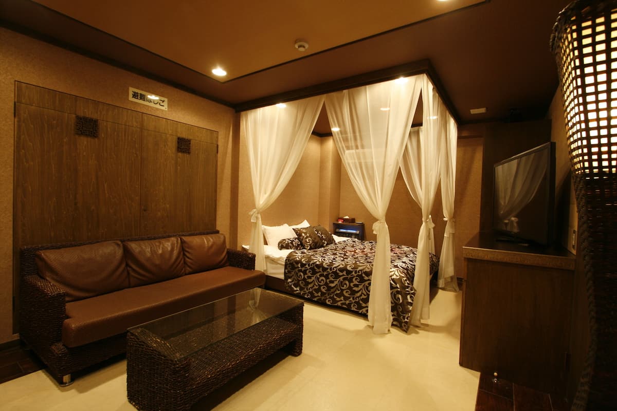 Ein Hotelzimmer mit einem braunen Ledersofa, einem Glastisch, einem Bett mit Baldachin und einem Fernseher auf einer Holzkassette.