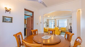 Zona pranzo con tavolo e sedie di legno, ventilatore a soffitto e vista su un soggiorno con divano giallo.