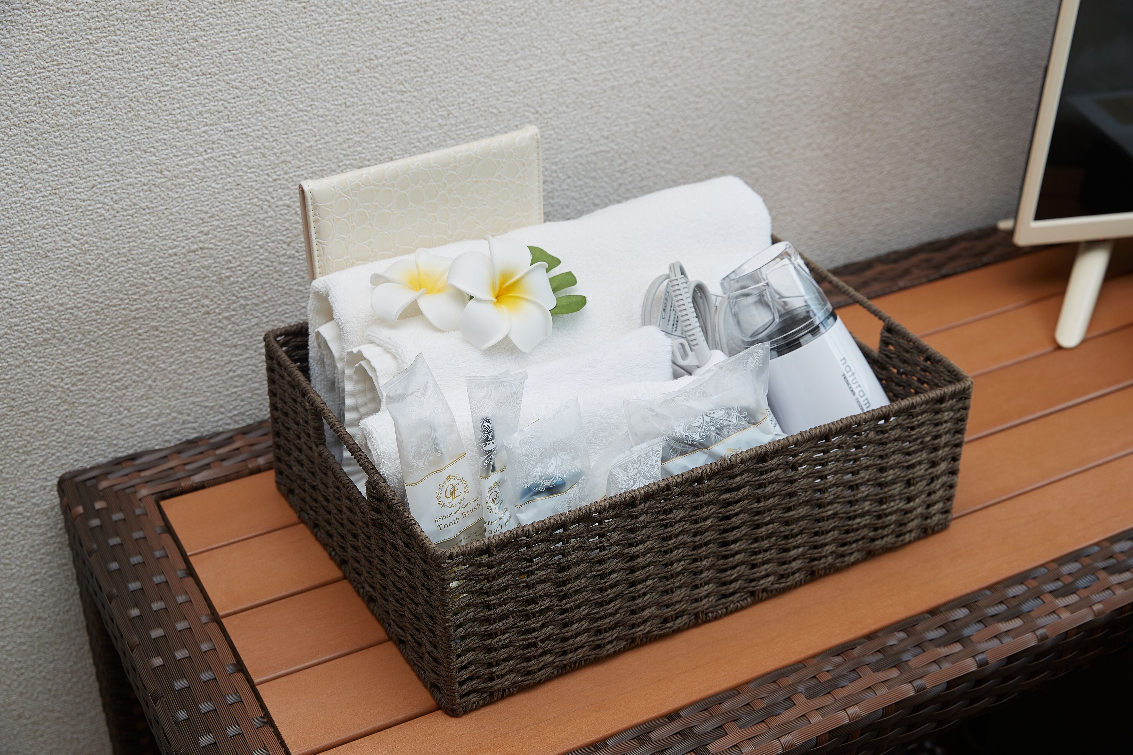 A wicker basket containing toiletries, a towel, and a flower on a wooden table.