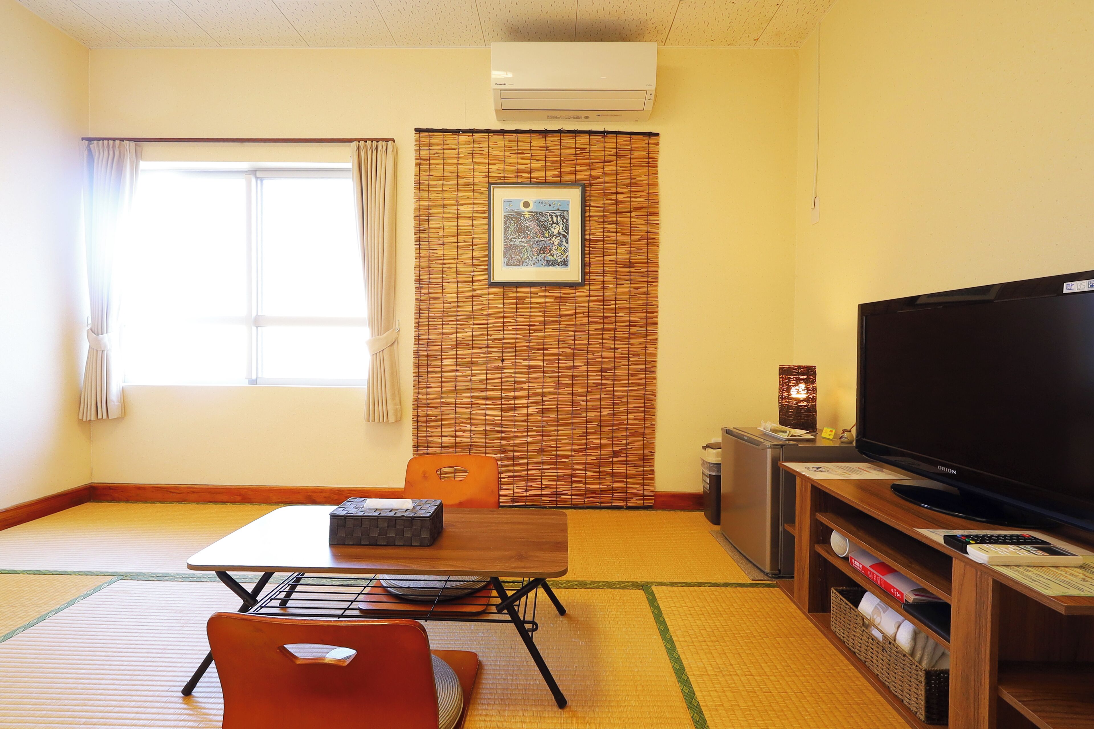 A room with tatami flooring, a television, a table with chairs, and a window with curtains.