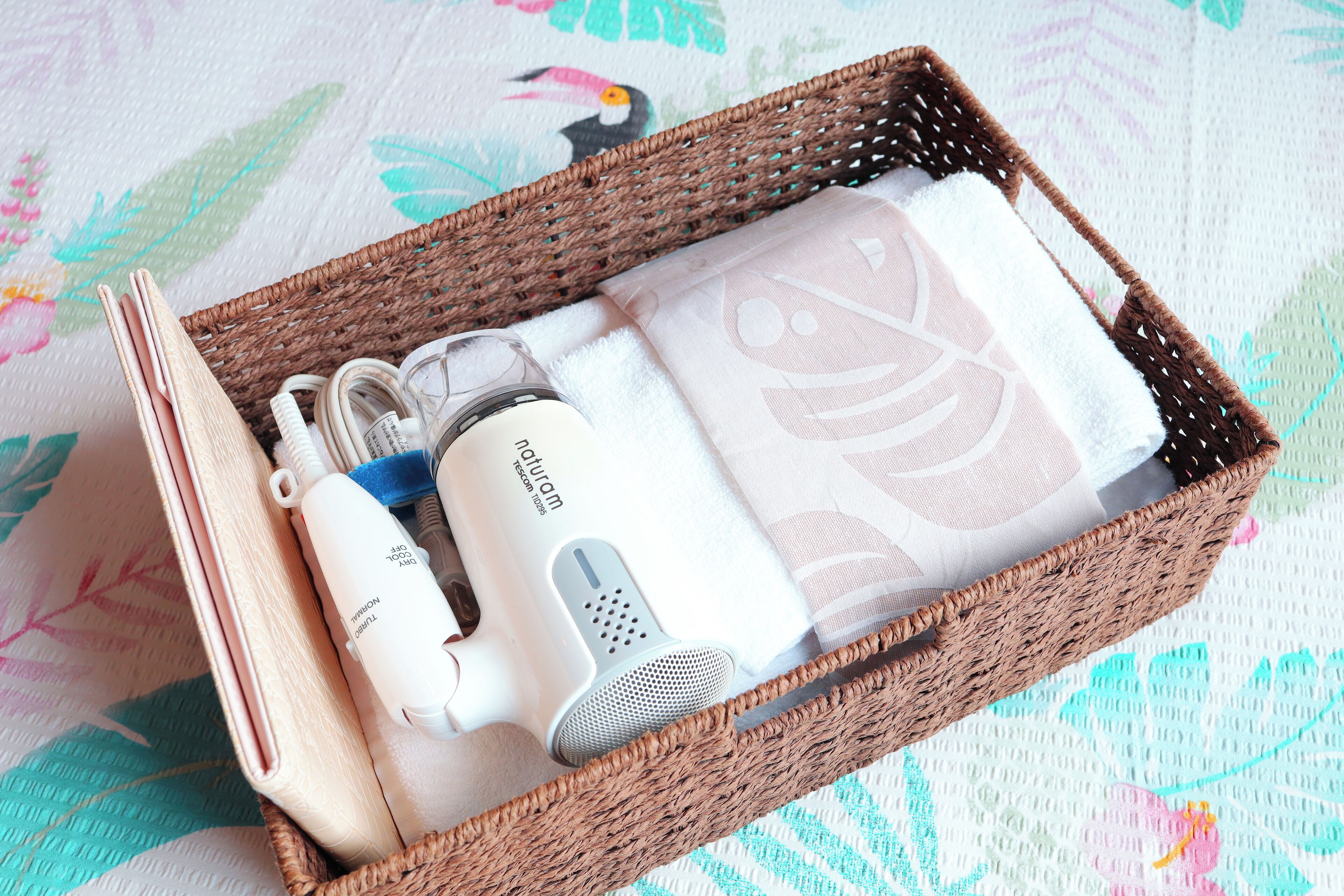 A wicker basket containing a hair dryer, a travel adapter, and a folded towel with a floral pattern.