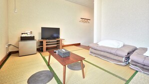 A room with tatami flooring, a television on a stand, a small refrigerator, and a bed with pillows. - Sazanka (Okinawa)