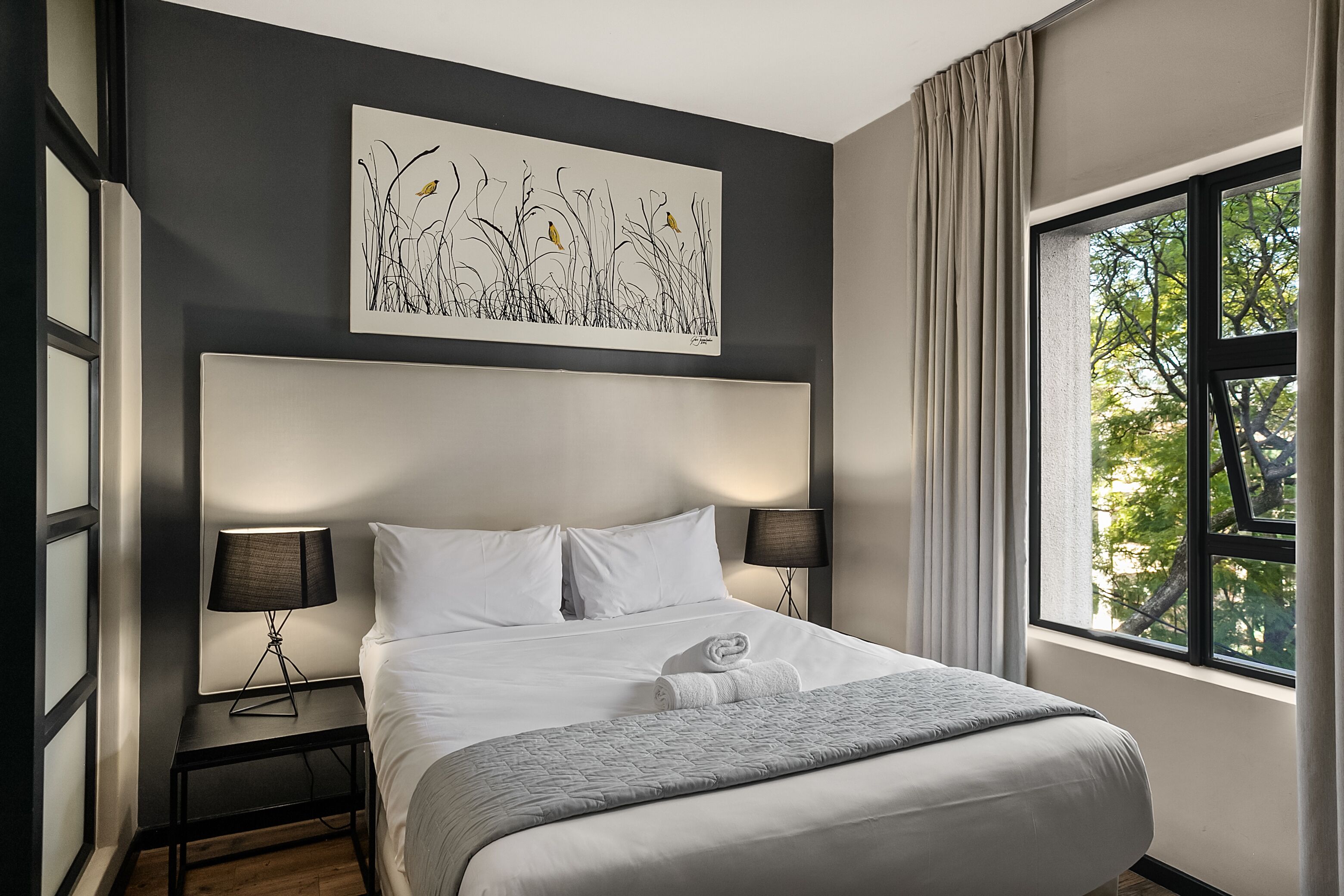A hotel room with a large bed, two bedside lamps, a window with curtains, and a wall-mounted artwork.