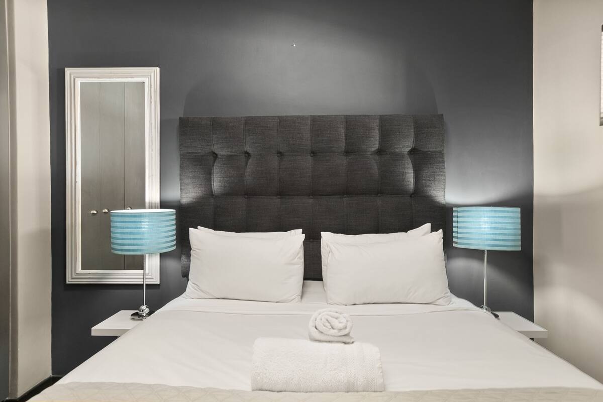 Una cama bien tendida con sábanas blancas, dos almohadas blancas y una toalla doblada sobre la cama. Hay dos mesitas de noche con lámparas, una a cada lado de la cama. La habitación tiene paredes oscuras y un cabecero capitonado.