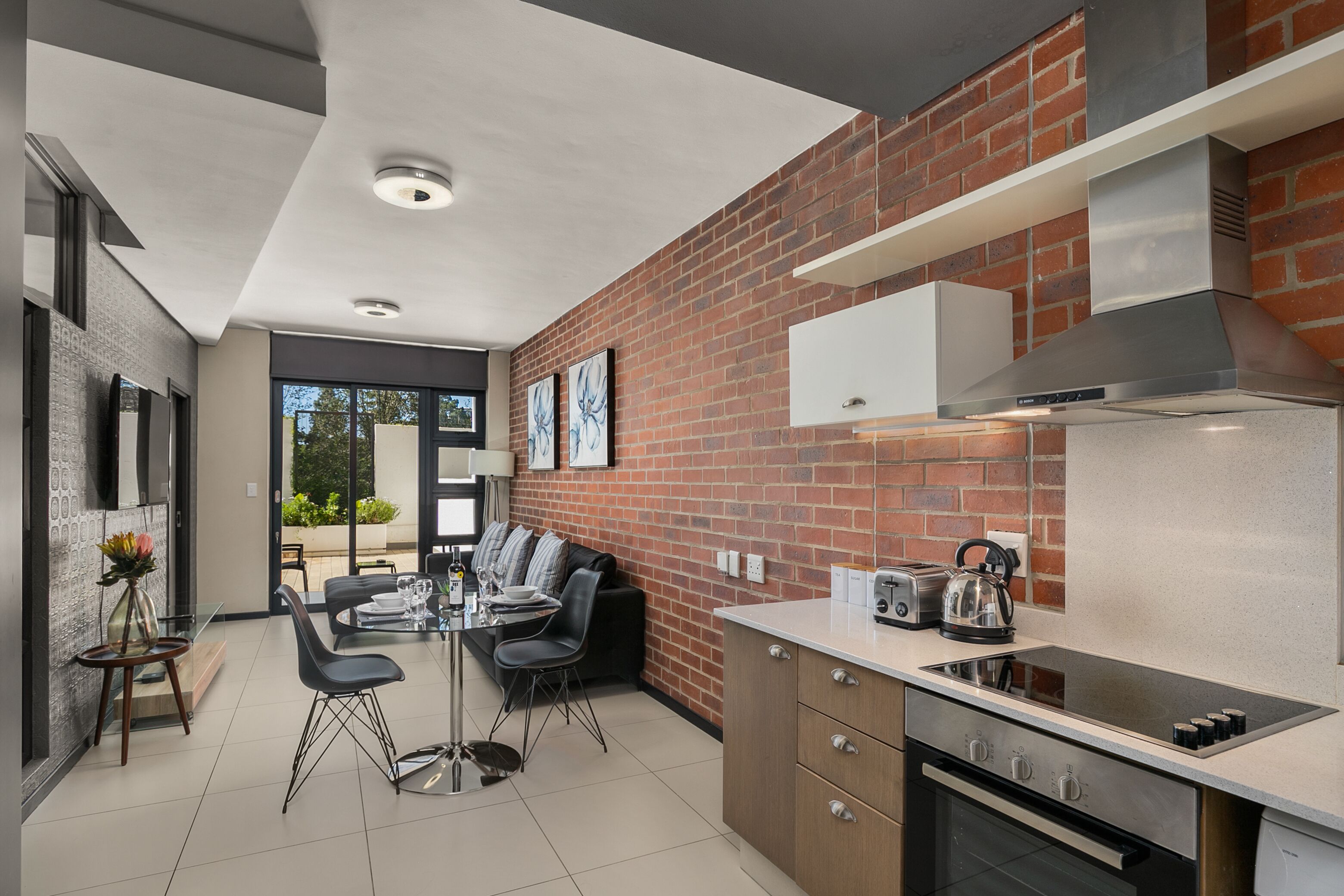 Een moderne keuken met een bakstenen muur, roestvrijstalen apparatuur en een eethoek met een glazen tafel en stoelen.