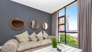 A modern living room with a grey wall, a sofa with patterned cushions, a potted plant, and a large window with a view of buildings and trees. - Easy Stay - The Vantage (Johannesburg)