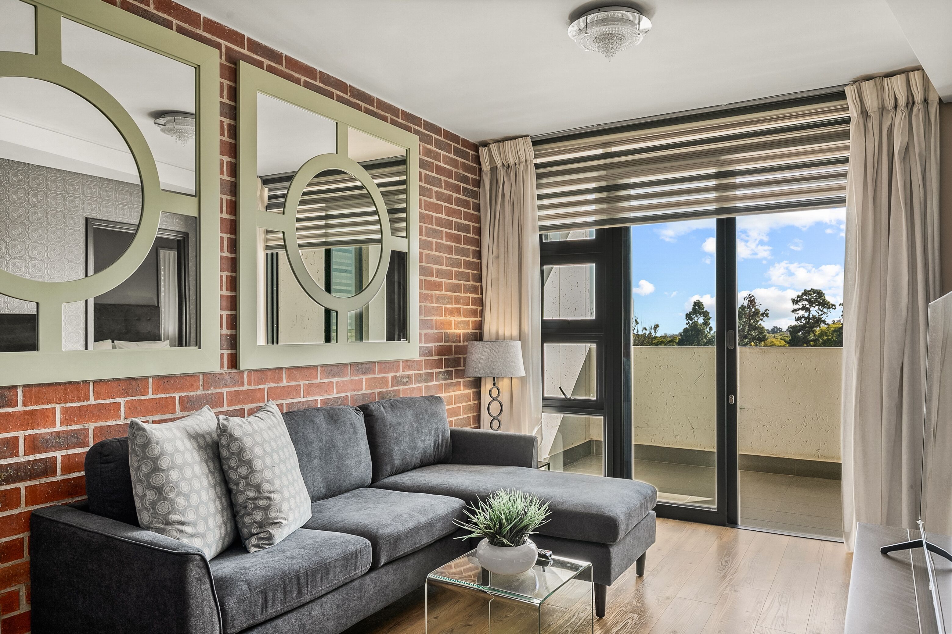 A modern living room with a grey sofa, a glass coffee table with a plant, and a large mirror on a brick wall.