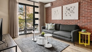 A modern living room with a grey sofa, a black chair, and three white round tables. There are two artworks on the brick wall and a small plant on a yellow side table. - Easy Stay - The Vantage (Johannesburg)