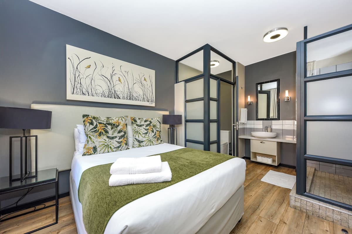 Una habitación de hotel moderna con una cama grande, un televisor de pantalla plana, un baño con espejo y lavamanos, y un cuadro en la pared.