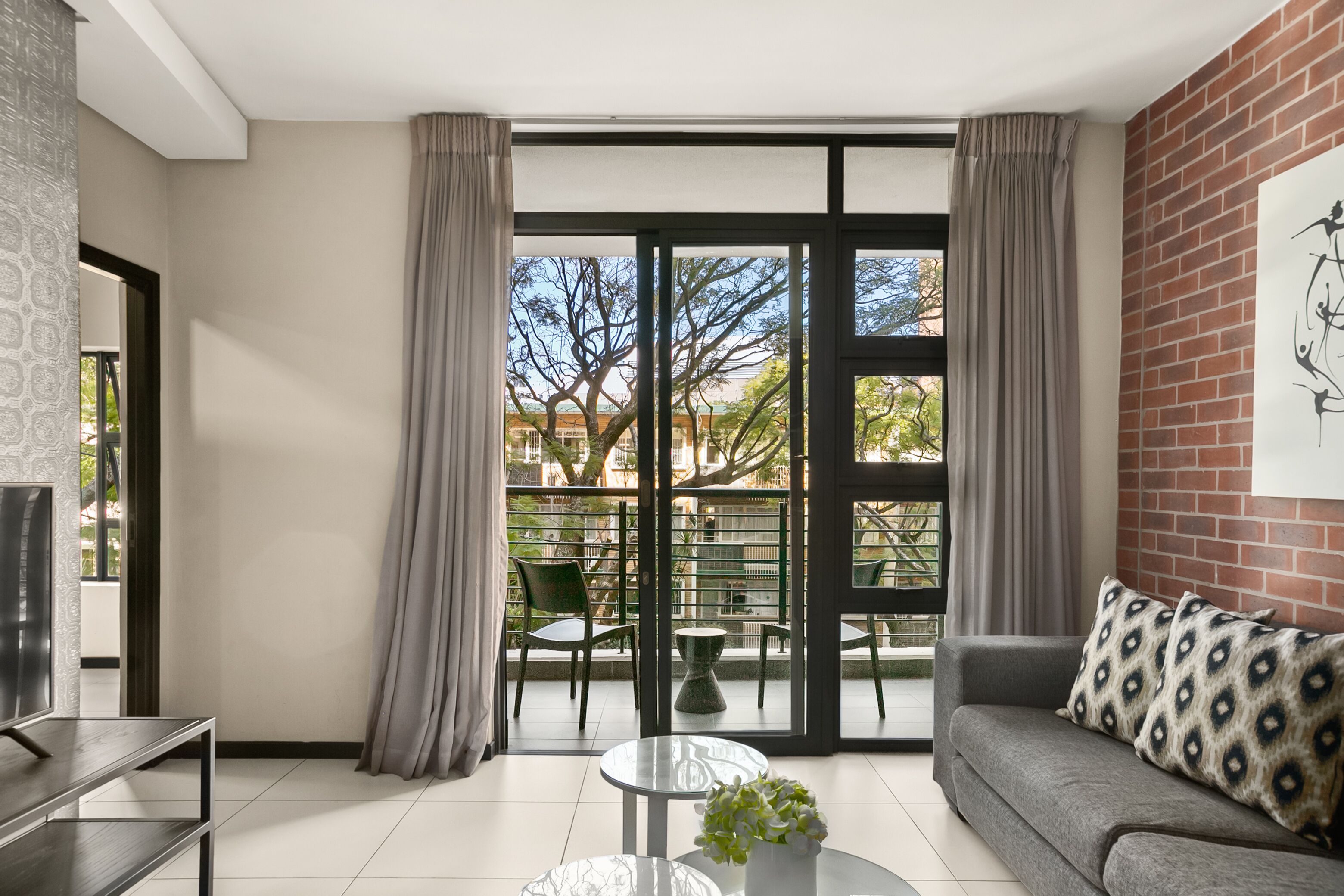 A modern living room with a grey sofa, a glass coffee table, and a TV on a stand. There is a balcony with a view of trees and buildings.