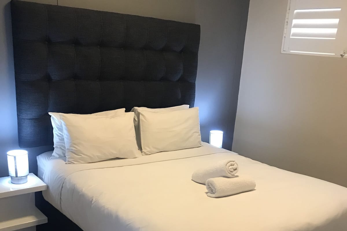 Una cama bien hecha con sábanas blancas y una toalla doblada, un cabecero tapizado oscuro y lámparas de noche sobre mesitas blancas.