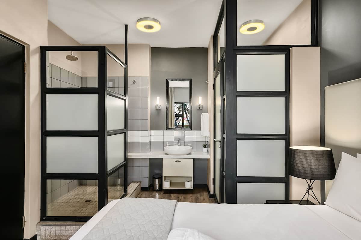 Una habitación de hotel moderna con una cama grande, una mesita de noche con lámpara, un baño con lavabo y espejo, y vistas al exterior a través de una puerta de cristal.