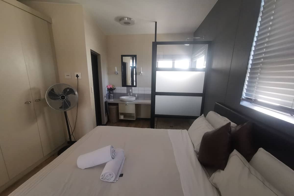 Habitación de hotel con cama, ventilador, baño con lavamanos y espejo, y ventana con persianas.