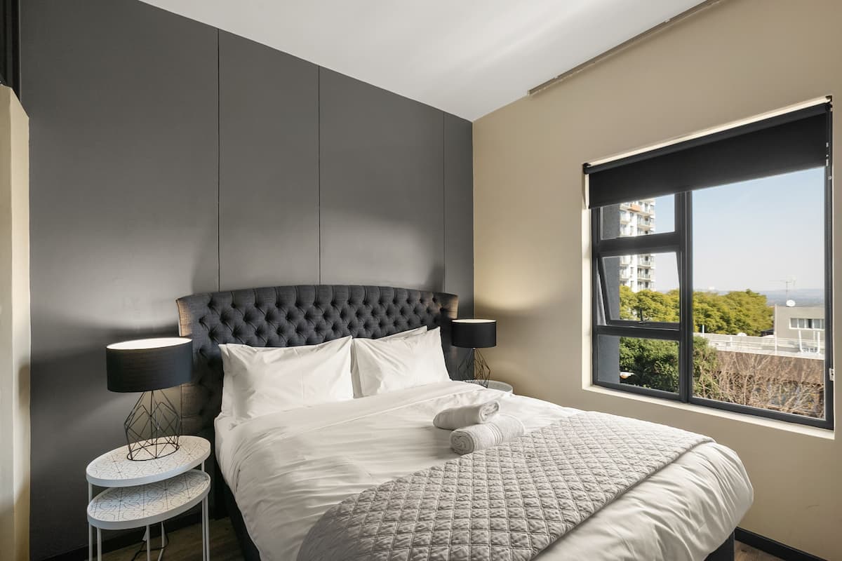 Un dormitorio moderno con cabecera capitonada, un ventanal con vistas a la ciudad, una mesita redonda con lámpara y cama bien tendida con toallas.