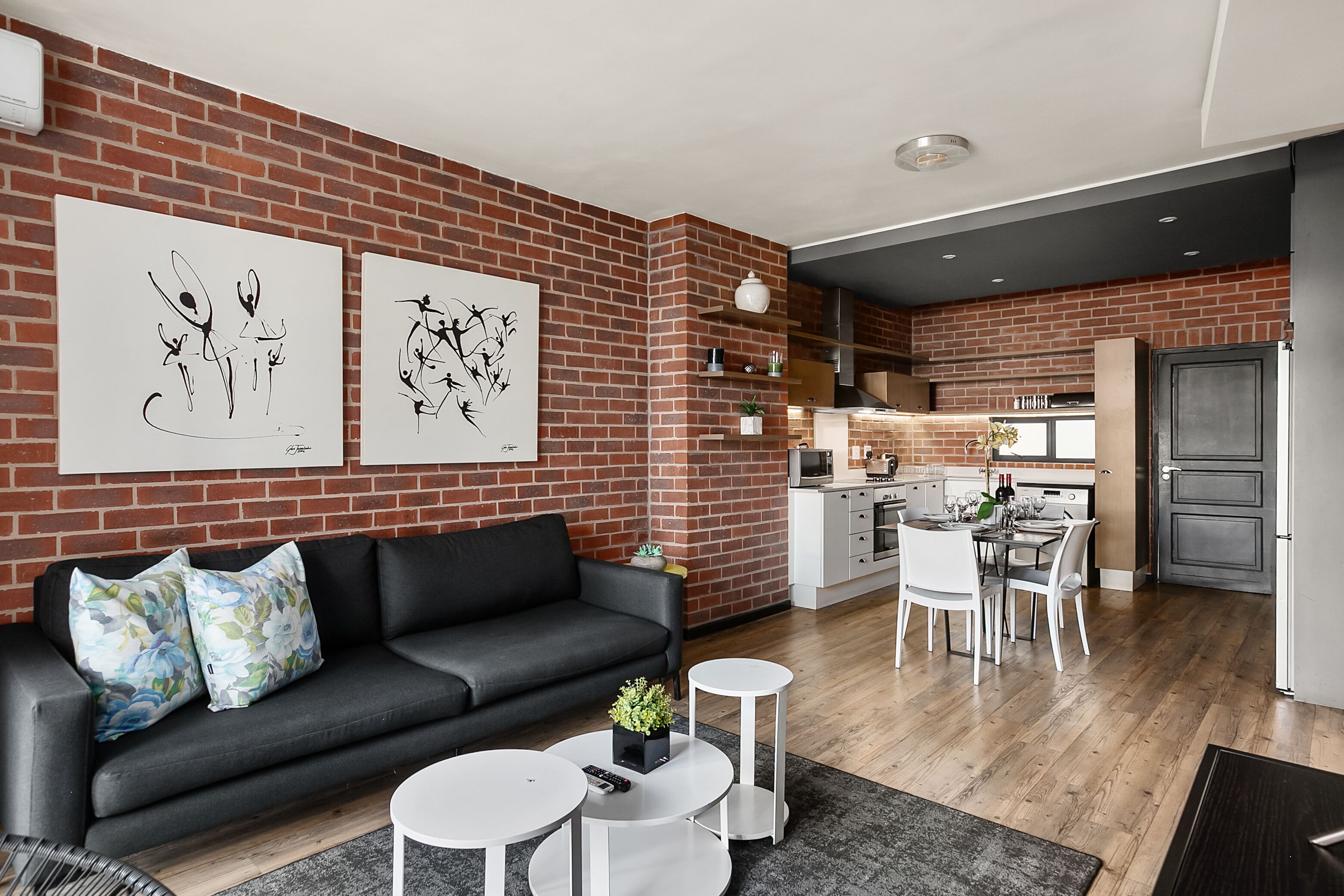 A modern living room with a black sofa, white coffee tables, and artwork on the brick wall.