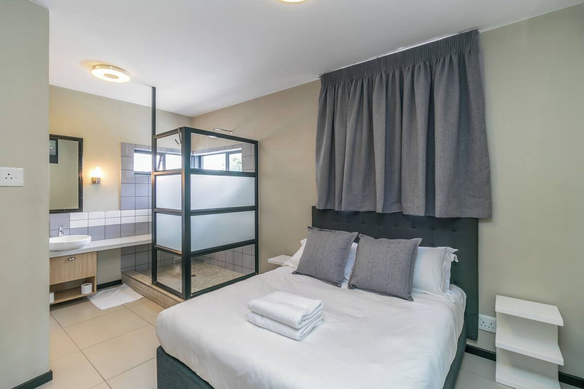 Una habitación de hotel moderna con una cama grande, cabina de ducha de vidrio, baño con lavamanos y espejo, y vista al exterior.