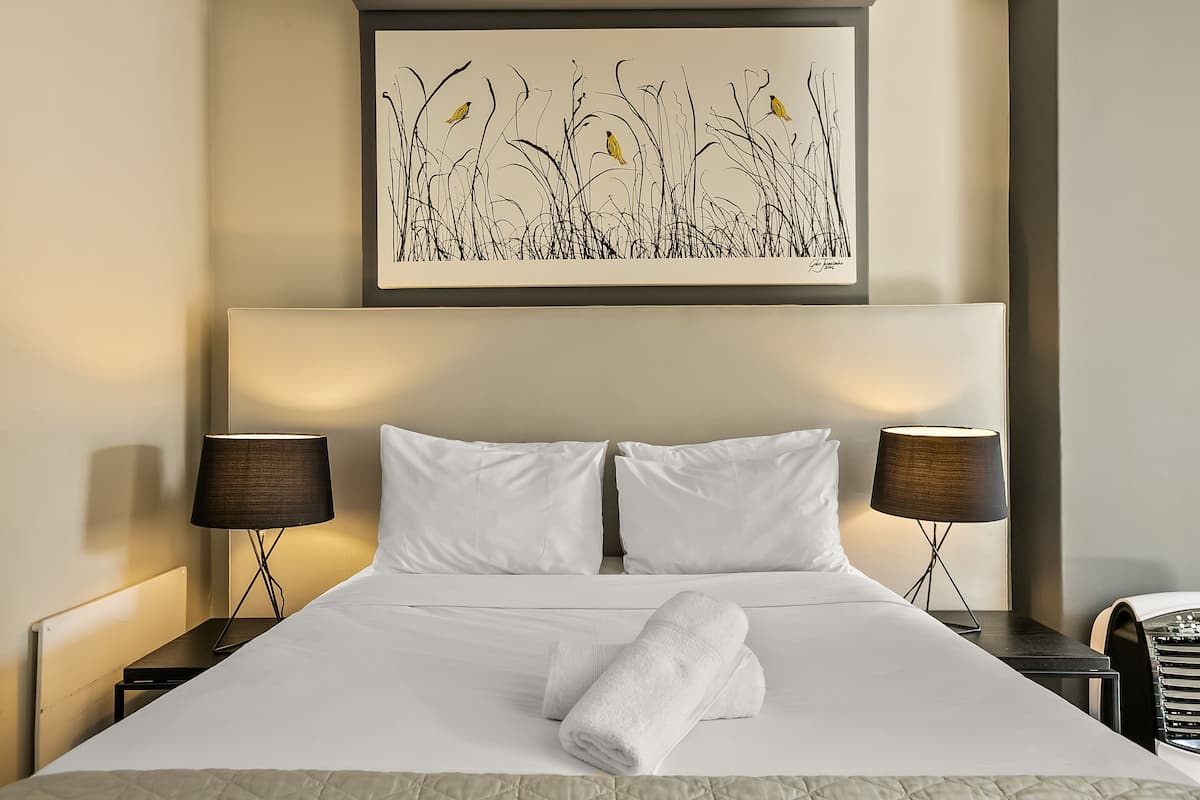 Una cama bien hecha con sábanas blancas, dos lámparas de noche y una obra enmarcada sobre la cama.