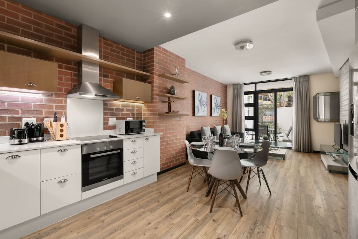 Una cocina moderna con armarios blancos, una pared de acento de ladrillo y un comedor con mesa y sillas de vidrio.