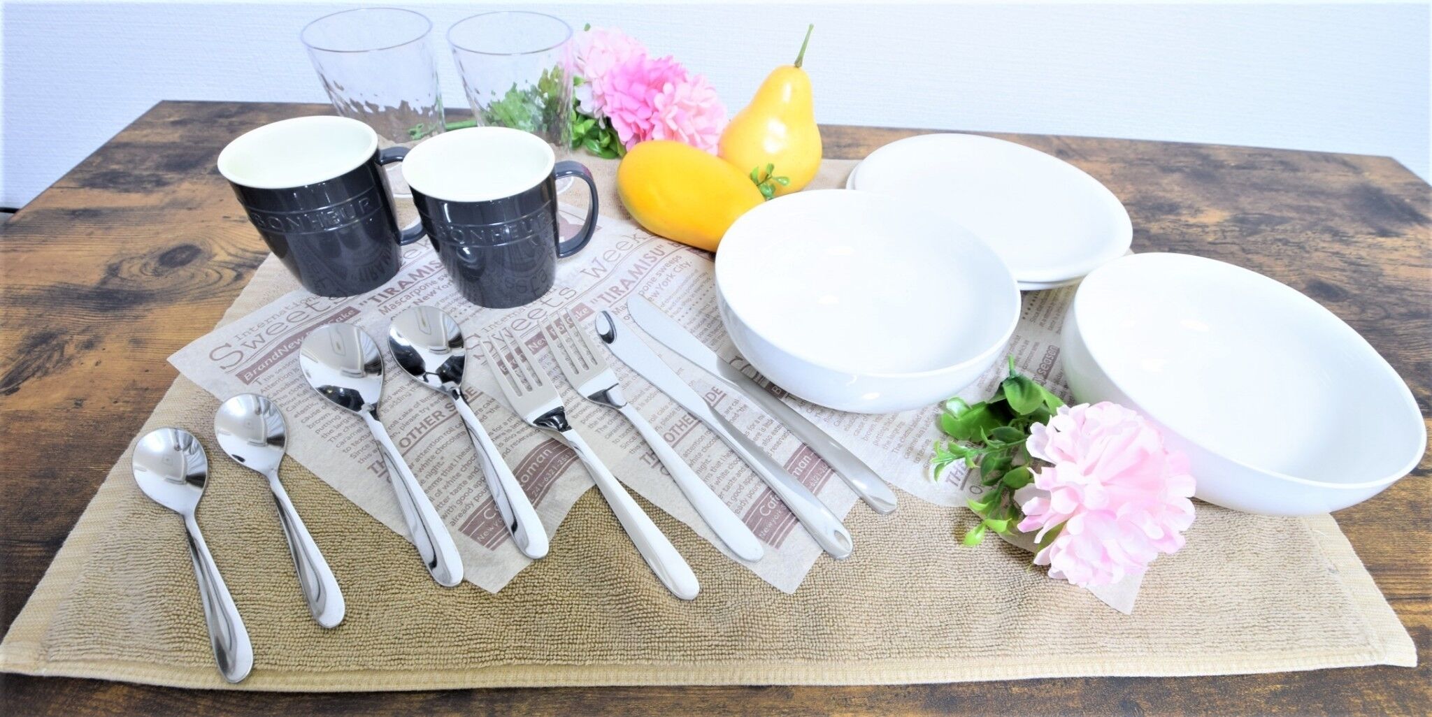 Une table dressée avec de l’argenterie, des assiettes et des bols blancs, le tout posé sur une nappe en toile de jute. On y trouve également un journal, des fleurs décoratives et une poire.