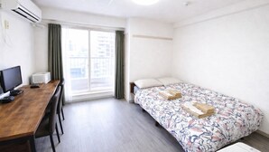 A hotel room with a wooden desk, a bed with floral bedding, a window with curtains, and an air conditioning unit. - Yado gh (Sapporo)