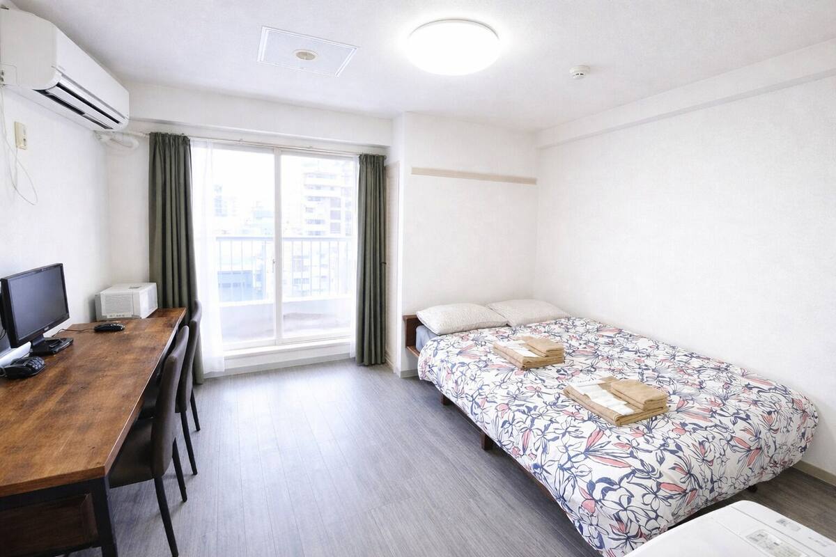 Une chambre d’hôtel avec un bureau en bois, un lit avec une literie à motifs floraux, une fenêtre avec des rideaux et un appareil de climatisation.